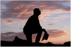 homme agenouillé dans la prière, tenant son chapeau à la main, sur un fond de ciel aux nuages rougeoyants à l’aurore ou au crépuscule